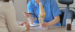 A doctor sits at a desk across from a patient, pointing to a model of a knee.